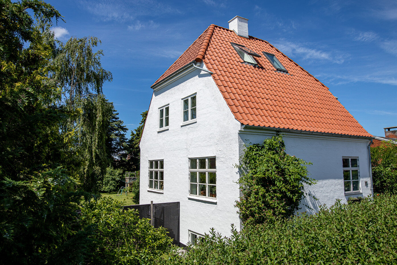 Hvidpudset murermestervilla med rødt tegltag og to ovenlysvinduer, omgivet af grønne træer